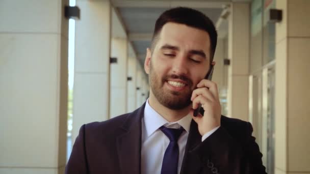 Close up view of smiling businessman dressed in shirt talking smartphone while walks on the background of a modern building — Stock Video