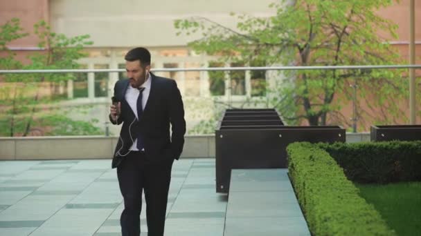 Felice uomo d'affari che ascolta musica in cuffia su smartphone, cammina fuori dall'ufficio e balla al ritmo . — Video Stock