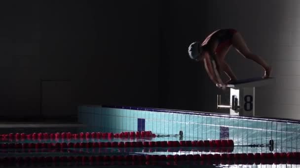 Мужчина-пловец прыгает с стартового камня и начинает плавать в бассейне. Профессиональная подготовка спортсменов: погружение и брызги поверхности воды. Ночной выстрел — стоковое видео