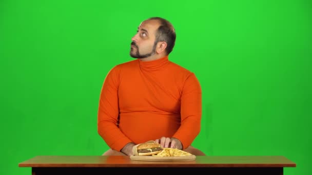 Чоловік з надмірною вагою дивиться на смачні нездорові продукти на столі і чинить опір нездоровій їжі, зелений екран — стокове відео