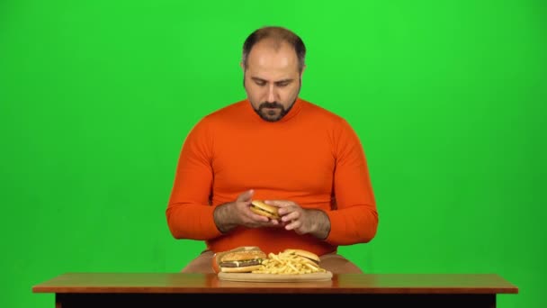 Blanke man aan tafel met bord fastfood kiest tussen hamburger en appel, stopt bij ongezond eten, groen scherm — Stockvideo