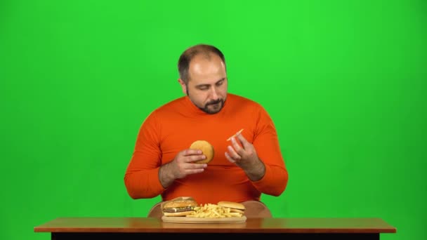 L'uomo a un tavolo con un piatto di fast food gode di mangiare un hamburger e patatine fritte, schermo verde — Video Stock