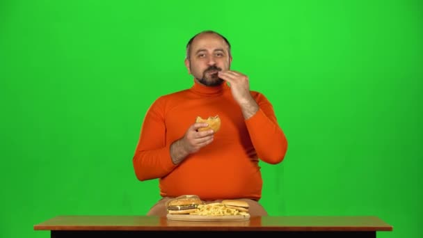 Hombre caucásico en una mesa con un plato de comida rápida disfruta comiendo una hamburguesa y papas fritas, pantalla verde — Vídeo de stock