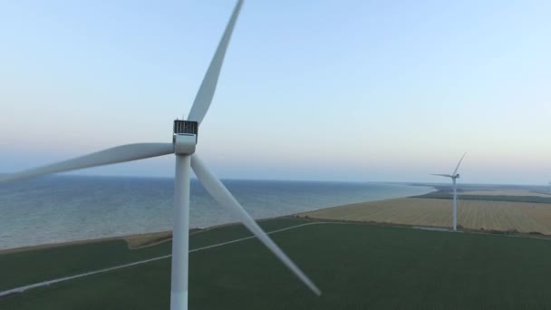 Molinos de viento a lo largo de la costa en campos verdes bajo cielos azules. Inspección aérea. De cerca. — Vídeo de stock