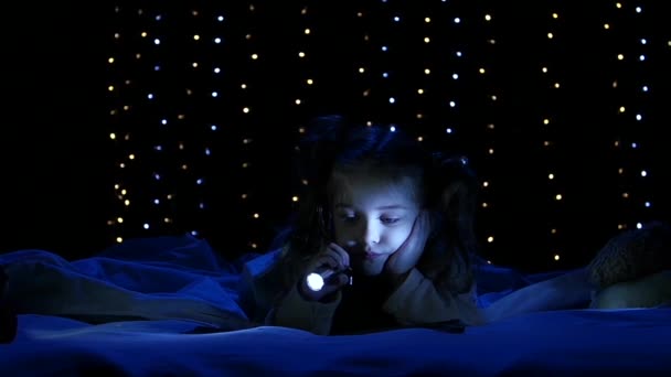 Egy kislány fekszik az ágyon, és zseblámpát ragyog a könyvre, amit olvas. Bokeh háttér. Lassú mozgás. — Stock videók