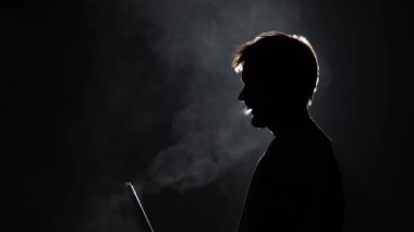 Profil görüntüsü silueti sakallı adam ağır çekimde siyah arka planda nargile içerken kalın duman üflüyor.