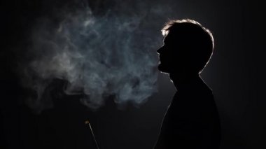 Profil görüntüsü silueti sakallı adam ağır çekimde siyah arka planda nargile içerken kalın duman üflüyor.