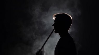 Profil görüntüsü silueti sakallı adam ağır çekimde siyah arka planda nargile içerken kalın duman üflüyor.