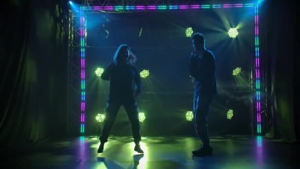 Silhouettes of a talented young girl and boy hip hop dancers. Hip hop street dance on a stage in dark studio with smoke and neon lighting. Dynamic lighting effects. Creative skills. Slow motion. — Stock Video