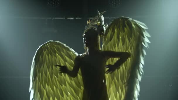 Силует танцюючої грецької богині Артеміди з крилами на сцені в темній студії з димом і неоновим освітленням. Яскрава молода жінка в золотій сукні і головному уборі. Динамічні світлові ефекти — стокове відео