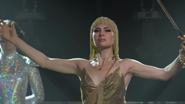 Dançando e cantando Joan of Arc no palco em um estúdio escuro com fumaça e iluminação de néon. Uma jovem brilhante com uma espada num vestido dourado e uma cobertura para a cabeça. Efeitos de iluminação de néon dinâmico. Luxuoso — Vídeo de Stock