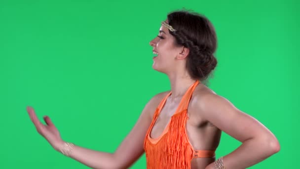 Portret van mooie jonge vrouwen zwaait koketterig met de hand en toont gebaar dat je hier komt. Een brandende brunette in een oranje franje op een groen scherm in de studio. Zijaanzicht. — Stockvideo