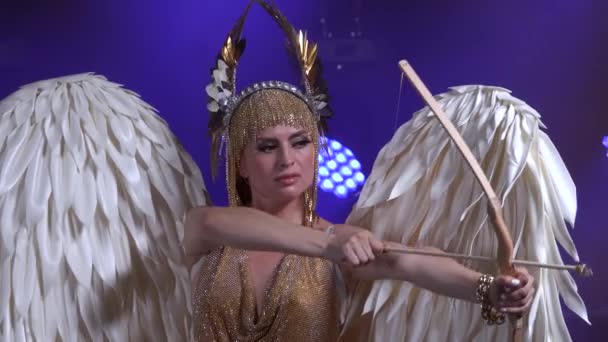 煙とネオンの照明と暗いスタジオでステージ上の弓と矢印と美しいギリシャの女神アルテミス。金色のドレスと頭飾りの翼を持つ明るい若い女性。ダイナミックなネオンライト — ストック動画
