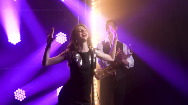 Uma linda vocalista cantando e gestos com as mãos no palco em um microfone vintage. No fundo é um saxofonista. Estúdio escuro com fumaça e iluminação de néon. Iluminação dinâmica de néon — Vídeo de Stock