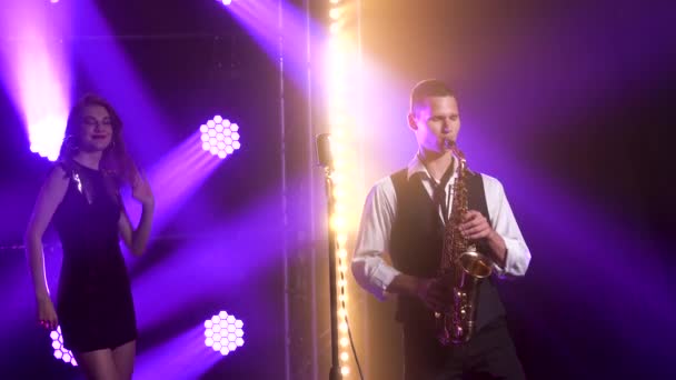 Um cara jovem e elegante toca o saxofone no palco e uma garota em um vestido preto com lantejoulas está dançando no fundo. Estúdio escuro com fumaça e iluminação de néon. Efeitos dinâmicos de iluminação de néon — Vídeo de Stock