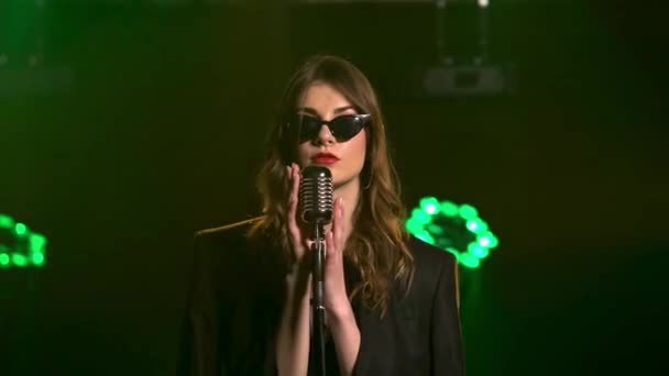 Una hermosa vocalista cantando lánguidamente y gestos con sus manos en el escenario en un micrófono vintage. Elegante en gafas retro negras y con lápiz labial rojo en los labios. Estudio oscuro con humo y — Vídeo de stock