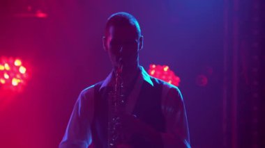 Siluet genç bir stil adamı sahnede saksafon çalıyor. Dumanlı ve neon ışıklı karanlık stüdyo. Dinamik neon ışık efektleri. Performans vokali ve müzik grubu. Kapat..