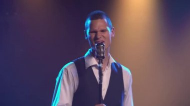 Genç stil sahibi bir vokalist elleri klasik bir mikrofonda sahnede şarkı söylüyor ve jestler yapıyor. Dumanlı ve neon ışıklı karanlık stüdyo. Dinamik neon ışık efektleri. Performans vokal ve