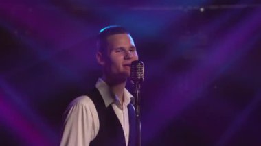 Genç stil sahibi bir vokalist duygusal olarak şarkı söylüyor ve elleri klasik bir mikrofonda sahnede hareketler yapıyor. Dumanlı ve neon ışıklı karanlık stüdyo. Dinamik neon ışık efektleri. Performans