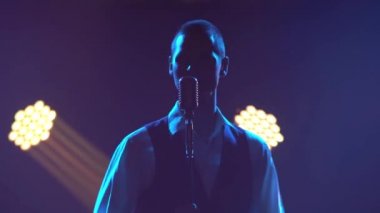Siluet, genç stil sahibi bir vokalist duygusal olarak şarkı söylüyor ve elleri klasik bir mikrofonda sahnede hareketler yapıyor. Dumanlı ve neon ışıklı karanlık stüdyo. Dinamik neon ışıklandırma efektleri