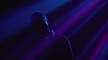 Siluet, genç stil sahibi bir vokalist duygusal olarak şarkı söylüyor ve elleri klasik bir mikrofonda sahnede hareketler yapıyor. Dumanlı ve neon ışıklı karanlık stüdyo. Dinamik neon ışıklandırma efektleri