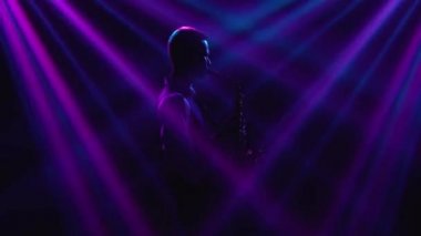Siluet genç ve şık bir adam sahnede parlak spot ışıklarıyla saksafon çalıyor. Dumanlı ve neon ışıklı karanlık stüdyo. Dinamik neon ışık efektleri. Performans vokali ve müzik grubu. Kenar