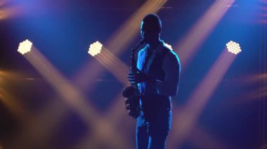 Siluet genç ve şık bir adam sahnede parlak spot ışıklarıyla saksafon çalıyor. Dumanlı ve neon ışıklı karanlık stüdyo. Dinamik neon ışık efektleri. Performans vokal ve müzik grubu.