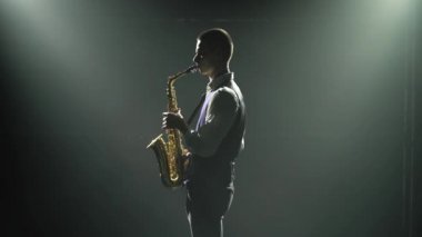 Siluet, genç ve şık bir adam sahnede altın gibi parlak bir saksafon çalıyor. Dumanlı ve neon ışıklı karanlık stüdyo. Performans vokali ve müzik grubu. Yan görünüm.