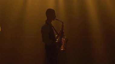 Siluet, genç ve şık bir adam sahnedeki sarı spot ışıklarında altın gibi parlak bir saksafon çalıyor. Dumanlı ve neon ışıklı karanlık stüdyo. Performans vokali ve müzik grubu. Yan görünüm.