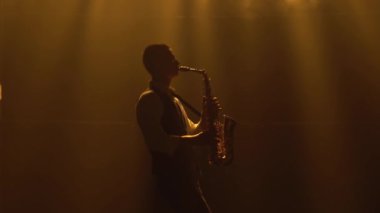 Siluet, genç ve şık bir adam sahnedeki sarı spot ışıklarında altın gibi parlak bir saksafon çalıyor. Dumanlı ve neon ışıklı karanlık stüdyo. Performans vokali ve müzik grubu. Yan görünüm.