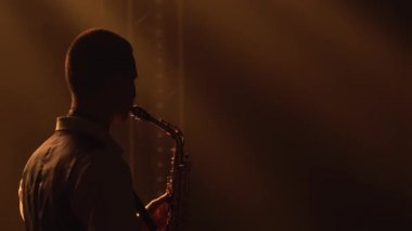 Siluet, genç ve şık bir adam sahnedeki sarı spot ışıklarında altın gibi parlak bir saksafon çalıyor. Dumanlı ve neon ışıklı karanlık stüdyo. Performans vokali ve müzik grubu. Arkadan bak. Kapat..