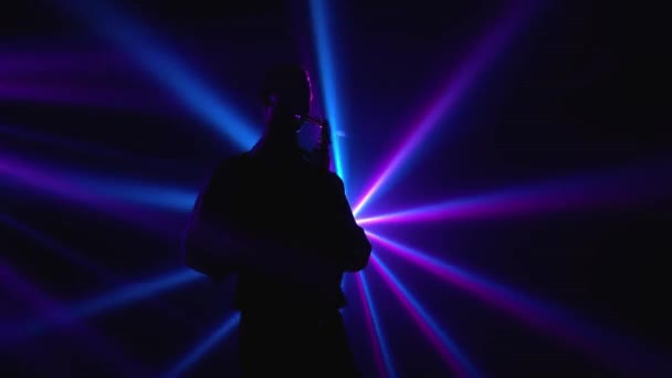 Silhouette, egy fiatal, stílusos fickó szaxofonozik a színpadon a fényes reflektorfényben. Sötét stúdió füsttel és neon világítással. Dinamikus neonfényhatások. Előadói ének és zenei zenekar. — Stock videók