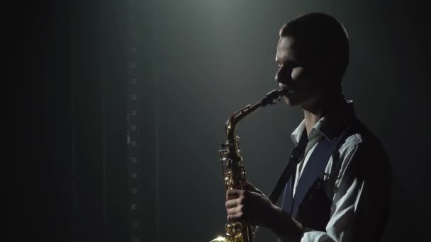 Silueta un chico joven y elegante toca el saxofón dorado brillante en el escenario. Estudio oscuro con humo e iluminación de neón. Banda vocal y musical. Vista lateral. De cerca.. — Vídeos de Stock