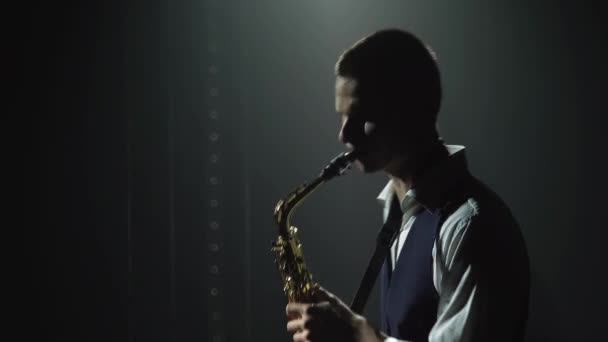 Silhueta um cara elegante jovem toca o saxofone dourado brilhante no palco. Estúdio escuro com fumaça e iluminação de néon. Performance vocal e banda musical. Vista lateral. Fechar . — Vídeo de Stock