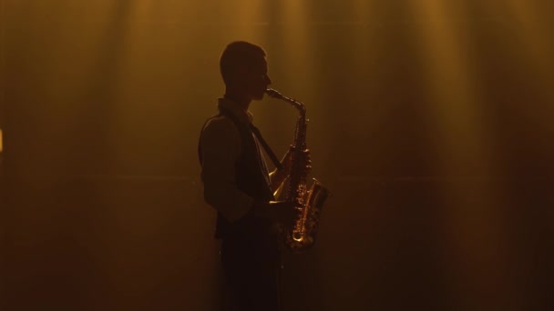 Silhouette spielt ein junger, stylischer Typ das goldglänzende Saxofon im gelben Scheinwerferlicht auf der Bühne. Dunkles Studio mit Rauch und Neonlicht. Performance Vokal- und Musikband. Seitenansicht. — Stockvideo