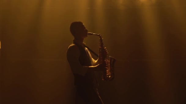 Silueta mladý stylový chlap hraje zlatý lesklý saxofon ve žlutých reflektorech na jevišti. Tmavé studio s kouřem a neonovým osvětlením. Vystupující vokální a hudební kapela. Boční pohled. — Stock video