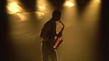 Siluet, genç ve şık bir adam sahnedeki sarı spot ışıklarında altın gibi parlak bir saksafon çalıyor. Dumanlı ve neon ışıklı karanlık stüdyo. Performans vokal ve müzik grubu.