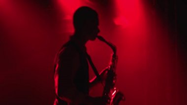 Siluet genç ve şık bir adam sahnedeki çok renkli spot ışıklarında altın gibi parlak bir saksafon çalıyor. Dumanlı ve neon ışıklı karanlık stüdyo. Dinamik neon ışık efektleri. Kapatın. Yan görünüm.