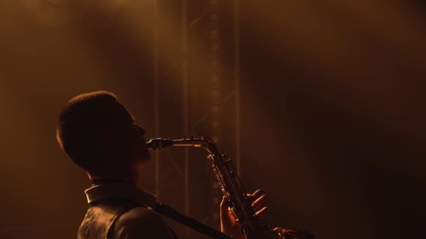 Silueta ayoung stylový chlap hraje zlatý lesklý saxofon ve žlutých reflektorech na jevišti. Tmavé studio s kouřem a neonovým osvětlením. Vystupující vokální a hudební kapela. Zadní pohled. Zavřít. — Stock video