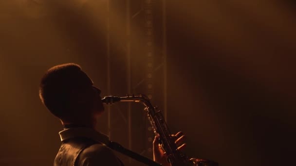 Silhouette spielt im gelben Scheinwerferlicht auf der Bühne das goldglänzende Saxofon. Dunkles Studio mit Rauch und Neonlicht. Performance Vokal- und Musikband. Rückansicht. Nahaufnahme. — Stockvideo