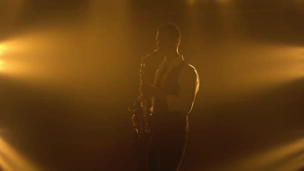 Silhouette un jeune homme élégant joue du saxophone doré brillant sous les projecteurs jaunes sur scène. Studio sombre avec fumée et néon. Performance groupe vocal et musical. — Video