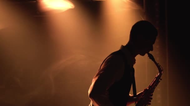 Silhouette un jeune homme élégant joue du saxophone doré brillant sous les projecteurs jaunes sur scène. Studio sombre avec fumée et néon. Accent sur le saxophone. Vue latérale. Gros plan. — Video