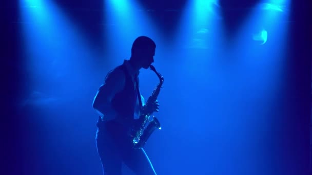 Silhouette spielt ein junger, stylischer Typ das goldglänzende Saxofon in den bunten Scheinwerfern auf der Bühne. Dunkles Studio mit Rauch und Neonlicht. Dynamische Neonlichteffekte. — Stockvideo