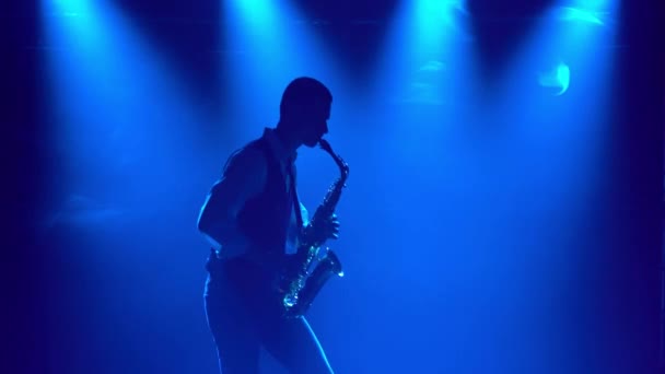 Silhueta um cara elegante jovem toca o saxofone dourado brilhante nos holofotes multicoloridos no palco. Estúdio escuro com fumaça e iluminação de néon. Efeitos dinâmicos de iluminação de néon . — Vídeo de Stock