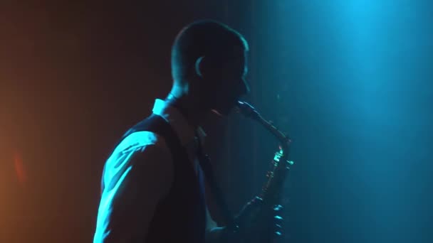 Silhouette un jeune homme élégant rétro joue lente mélodie sur le saxophone doré brillant sur scène. Studio sombre avec fumée et néon. Gros plan. — Video