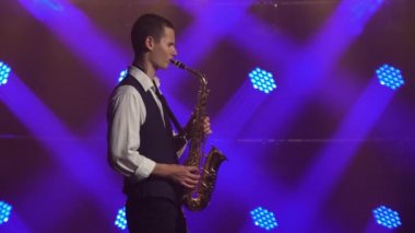 Genç, modaya uygun bir adam sahnedeki mavi spot ışıkları altında altın renkli saksafonda çalıyor. Dumanlı ve neon ışıklı karanlık stüdyo. Yan görünüm.