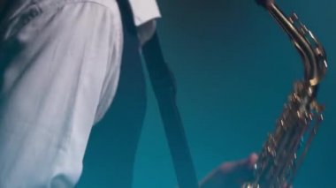 Genç, modaya uygun bir adam, sahnedeki turkuaz spot ışıklarıyla altın gibi parlak bir saksafonda çalar. Dumanlı ve neon ışıklı karanlık stüdyo. Eller ve saksafon yakın çekim. Yan görünüm.