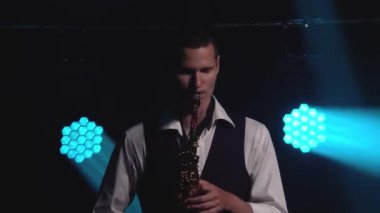 Genç, modaya uygun bir adam, sahnedeki turkuaz spot ışıklarıyla altın gibi parlak bir saksafonda çalar. Dumanlı ve neon ışıklı karanlık stüdyo. Kapat..