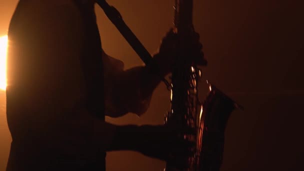 Silhouette un jeune homme élégant joue du saxophone doré brillant sous les projecteurs jaunes sur scène. Studio sombre avec fumée et néon. Accent sur le saxophone. Gros plan. — Video