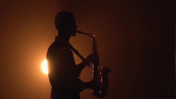 Silhouette spielt ein junger, stylischer Typ das goldglänzende Saxofon im gelben Scheinwerferlicht auf der Bühne. Dunkles Studio mit Rauch und Neonlicht. Akzent auf dem Saxofon. Seitenansicht. Nahaufnahme. — Stockvideo